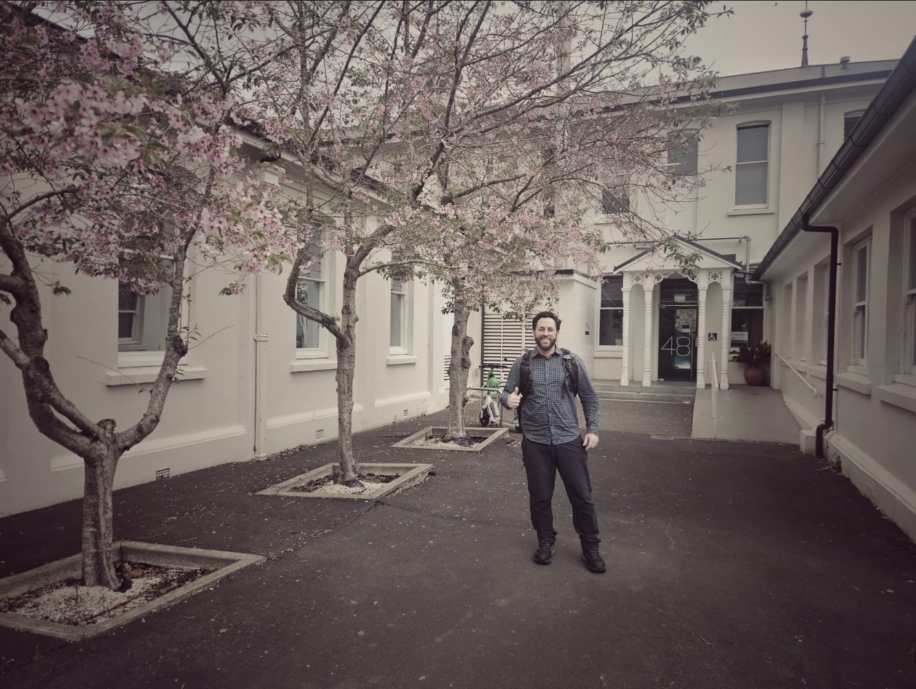 Thiago Amaral giving a thumbs up in front of Building 48 at Unitec, under blossoming cherry trees